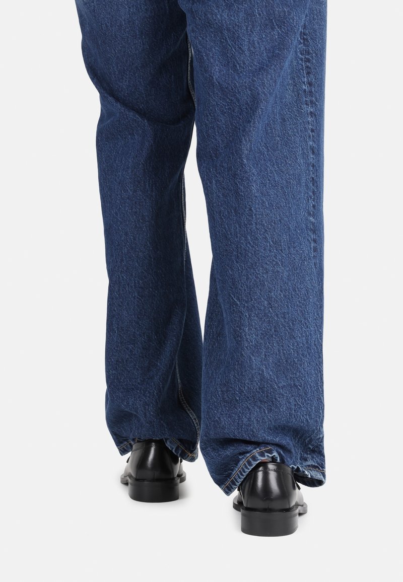 Jean bleu en denim à jambes larges avec une texture délavée, associés à des chaussures en cuir noires avec une semelle robuste et un bout arrondi.