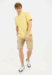 Geel kortgeleden overhemd met knopen en een borstzak, gecombineerd met beige cargoshorts. Model draagt witte sneakers met zwarte accenten.