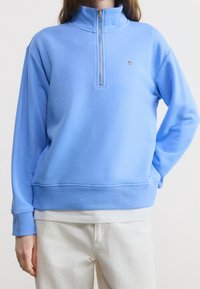 Femme portant un sweat-shirt à fermeture éclair partielle bleu clair et un pantalon blanc, debout avec une main dans la poche sur un fond uni.