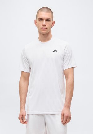 Mężczyzna z krótkimi włosami, ubrany w zwykły biały t-shirt Adidas i białe spodenki, stojący na jasnoszarym tle.