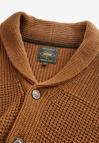 Brązowy dziergany sweter rozpinany z fakturowanym splotem, kołnierzem typu szalowego, dwoma guzikami oraz metką z napisem „Premium Grade 1982 NEXT Durable Goods.”