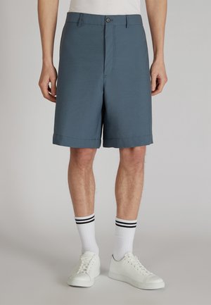 Pantaloni blu con una texture liscia, un taglio dritto e un orlo piegato, abbinati a sneakers bianche e calze sportive a righe.