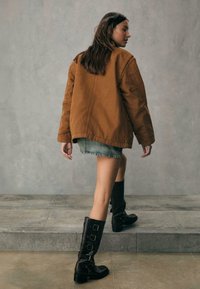 Brązowa, oversize'owa kurtka, dżinsowa mini spódnica i czarne skórzane kozaki za kolano z klamrami. Modelka idzie w kierunku alejki na kamiennej nawierzchni.