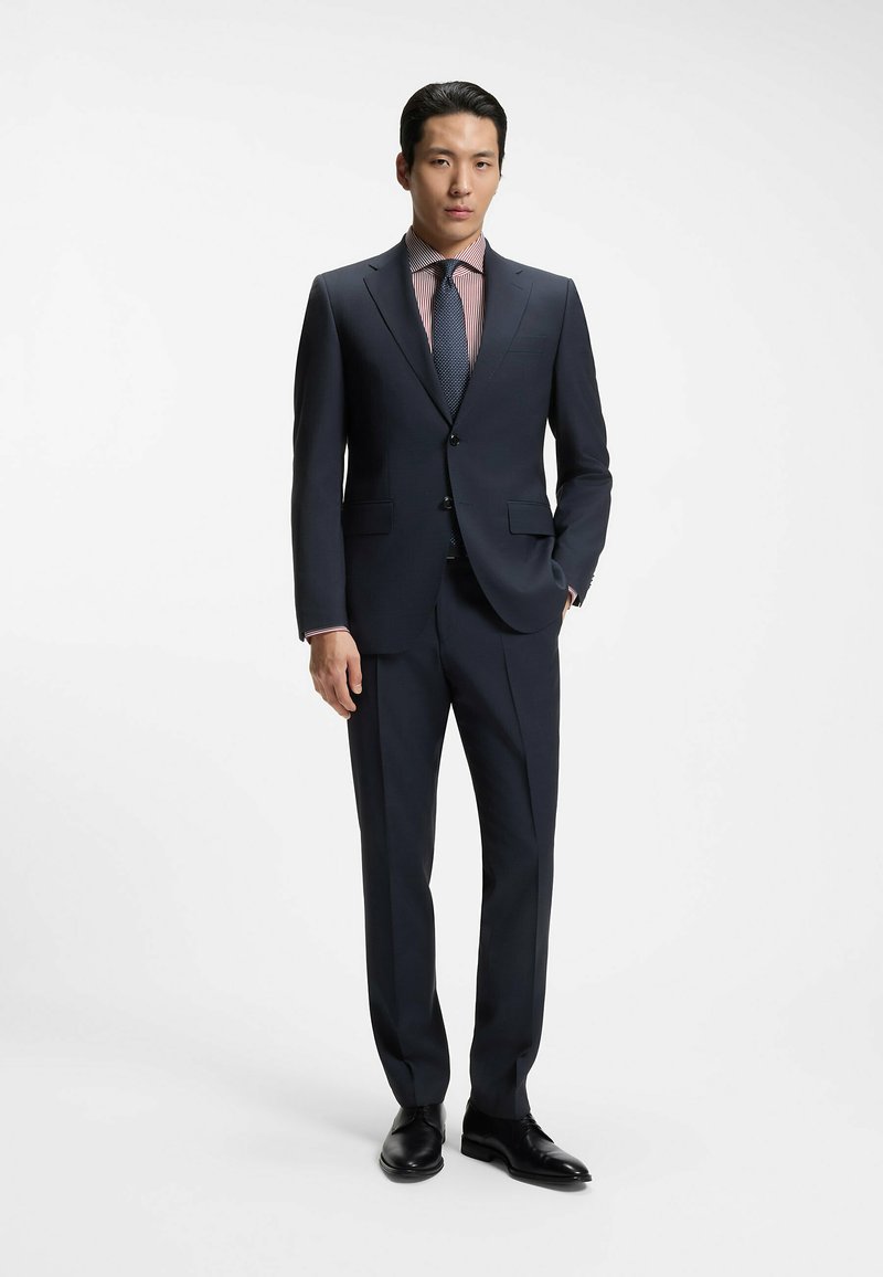Homme portant un costume bleu marine foncé avec des chaussures noires, une chemise rayée rouge et blanche, et une cravate sombre à motifs sur un fond blanc uni.