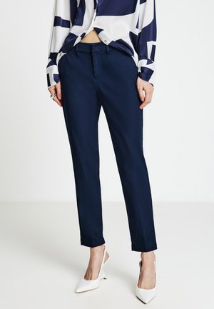 Donna che indossa pantaloni su misura blu navy, tacchi bianchi a punta e una camicetta con motivo blu navy e bianco con un bottone slacciato.