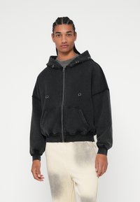 Crna oversized zip-hodnik izrađena od mekanog materijala, s kapuljačom, prednjim džepovima i manžetama. Teksturalna završnica s zaobljenim rubom.