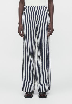 Personne portant un pantalon taille haute, à jambes larges, avec des rayures verticales bleu marine et blanches et des chaussures noires sur un fond uni.