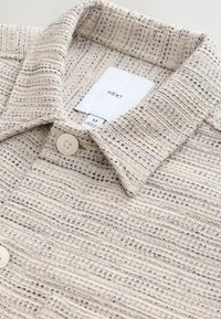 Chemise beige texturée à boutons avec col classique, boutons crème et étiquette de marque "NEXT" visible à l'intérieur de l'encolure.