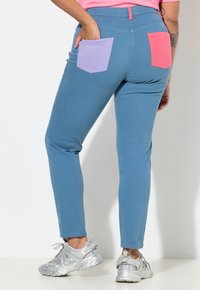 Modré kalhoty s úzkým střihem, na zadní straně mají fialové a korálové nášivkové kapsy. Látka má hladkou texturu. Kombinovány se stříbrnými teniskami.