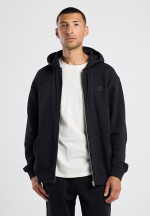 Zwarte rits-hoodie van zachte stof met een capuchon, voorzakken en een klein logo-detail op de borst. Draag het over een licht shirt.