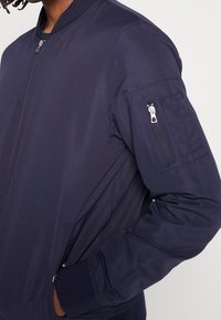 Granatowa kurtka bombowa o gładkiej fakturze, z zamkiem z przodu, prążkowanymi mankietami i zapinaną na zamek kieszenią na lewym rękawie.