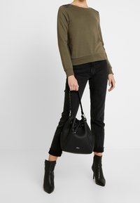 Sudadera verde oliva con acentos texturizados en los hombros, combinada con jeans negros, botas de tobillo de tacón y un bolso de cubo negro.