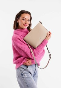 Béžová kožená kabelka s elegantním designem, vybavená horním zipem a nastavitelným ramenním popruhem, kterou drží osoba v růžovém svetr.