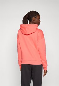 Korallfärgad hoodie med dragsko, avslappnad passform, mjukt tyg och ribbade cuffs. Kombinerad med mörka, löst sittande byxor. Bakvy.