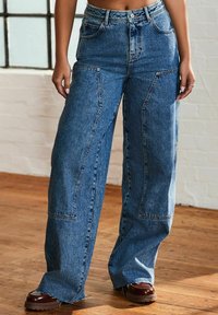 Jeans largi din denim albastru, cu talie înaltă, având buzunare frontale mari, cusături în contrast și un tiv brut la manșete.