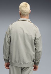 Lys grå jakke med et struktureret stof, der har krave, elastiske manchetter og et lille logo-patch nær sømmen. Enkel design.