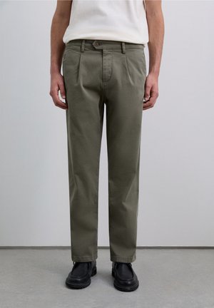 Bărbat îmbrăcat cu pantaloni plisati de culoare verde măsliniu, încălțat cu pantofi negri, și purtând o cămașă albă băgată în pantaloni, stând pe un podea gri, lângă un perete simplu.