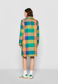 Abrigo de cuadros colorido con tonos verdes, naranjas, amarillos y azules, con una silueta ajustada y mangas largas, combinado con calcetas blancas y zapatillas acentuadas en verde.