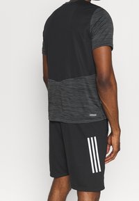 Mies, joka on pukeutunut musta-harmaaseen lyhythihaiseen urheilupaitaan ja mustiin shortseihin, joissa on kolme valkoista raitaa sivussa, kääntyy poispäin.