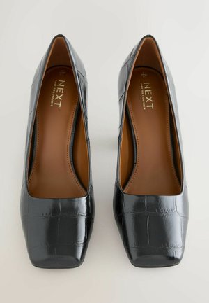 Mustad lakknahast kõrge kontsaga kingad ruudukujulise varbaga. Tekstuuritud viimistlus, millel on krokodillinahk muster; sisemus on pruun ja bränditud.