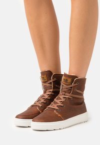 Brown leather high-top sneakers with light stitching, brown laces, padded collar, and white rubber sole. Hub logo on the tongue.