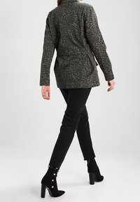Chaqueta de estampado de leopardo en verde oscuro y negro, con una textura suave, mangas largas y cintura ceñida. Combinada con jeans ajustados negros y botines.