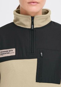 Fleece- og stoff halv-zip genser i svart og beige, med en glidelåslomme på brystet og merker med "Designet med dedikasjon."