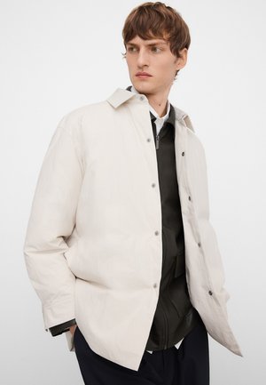 Junger Mann trägt ein geschichtetes Outfit mit einer hellbeigen, übergroßen Jacke über einer dunklen Reißverschlussjacke, Hände in den Taschen, schaut nach links.