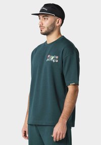Mørkegrønn oversized t-skjorte med korte ermer, rund hals, med flerfarget "PAS DE MONACO" tekst på brystet og et lite merke på ermet.