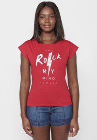 Camiseta roja de manga corta con el texto "Siempre Rockeas Mi Mente" y un gráfico de rayo; combinada con pantalones cortos de mezclilla. Textura suave.