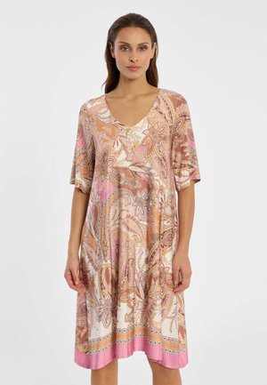 Frau in einem knielangen Kleid mit Paisley-Muster in Pink, Beige und Braun sowie ellbogenlangen Ärmeln, die vor einem schlichten Hintergrund steht.