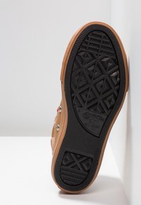 Zapato de cuero marrón claro con suela de goma negra, que presenta un patrón texturizado y acentos rojos. El logo de Converse All Star es visible en la suela.