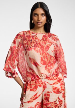 Kvinde med langt sort hår iført løs rødrandet og beige mønstret bluse og matchende bukser med vide ben, stående mod en ensfarvet baggrund.