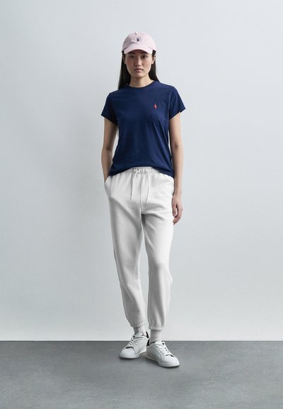 Νέα γυναίκα στέκεται, φορώντας μπλε σκούρο μπλουζάκι, ανοιχτό ροζ καπέλο, λευκή φόρμα και λευκά αθλητικά παπούτσια, με φόντο ένα απλό γκρι φόντο.