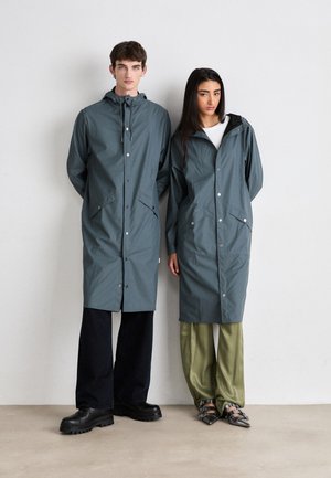Deux personnes portant des manteaux de pluie légers et longs de couleur turquoise avec des cordons, des boutons-pression et des poches avant. L'un porte un jean noir, l'autre un pantalon olive.