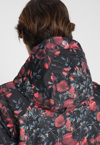 Schwarze Jacke mit Blumenmuster, die rote Mohnblumen zeigt, mit Kapuze und einem Logo auf dem Rücken. Das Material wirkt glatt und leicht.