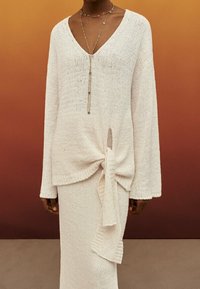Pull crème en tricot avec un col V, des manches longues et un détail de nœud sur le côté, associé à une jupe longue assortie avec un accent drapé.