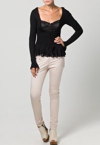 Nainen, jolla on yllään muotoiltu musta pitkähihanen paita, jossa on edessä vetoketju ja rusetti, beige kapea housut ja beige avokärkinen korkokenkä, seisoo käsi lantiolla.