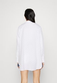 Camicetta bianca con texture a pois, maniche lunghe e vestibilità rilassata. La vista posteriore mostra il colletto e l'orlo curvo. Materiale leggero.