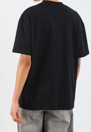 Osoba ubrana w gładki czarny t-shirt z krótkim rękawem i jasnoszare dżinsy, widoczna od tyłu na jednolitym białym tle.