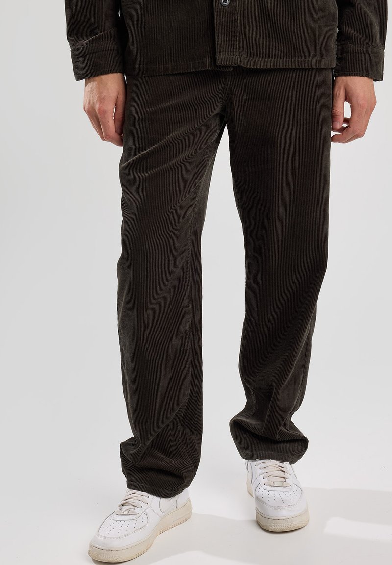 Dark green corduroy trousers with vertical stripes, loose fit, and a cuffed hem, paired with white sneakers.