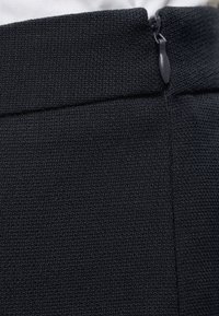 Gros plan sur la ceinture d'un pantalon en tissu noir texturé avec une petite tirette de fermeture éclair noire sur la couture latérale.