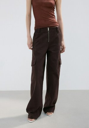 Femme portant un pantalon cargo large marron foncé avec fermeture éclair à l'avant et un top marron sans manches, debout sur un sol gris clair.