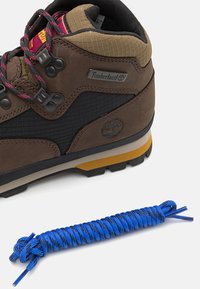 Scarponcino da trekking marrone con parte superiore testurizzata, accenti grigi e neri, e suola gialla, abbinato a un paracord blu coiled.