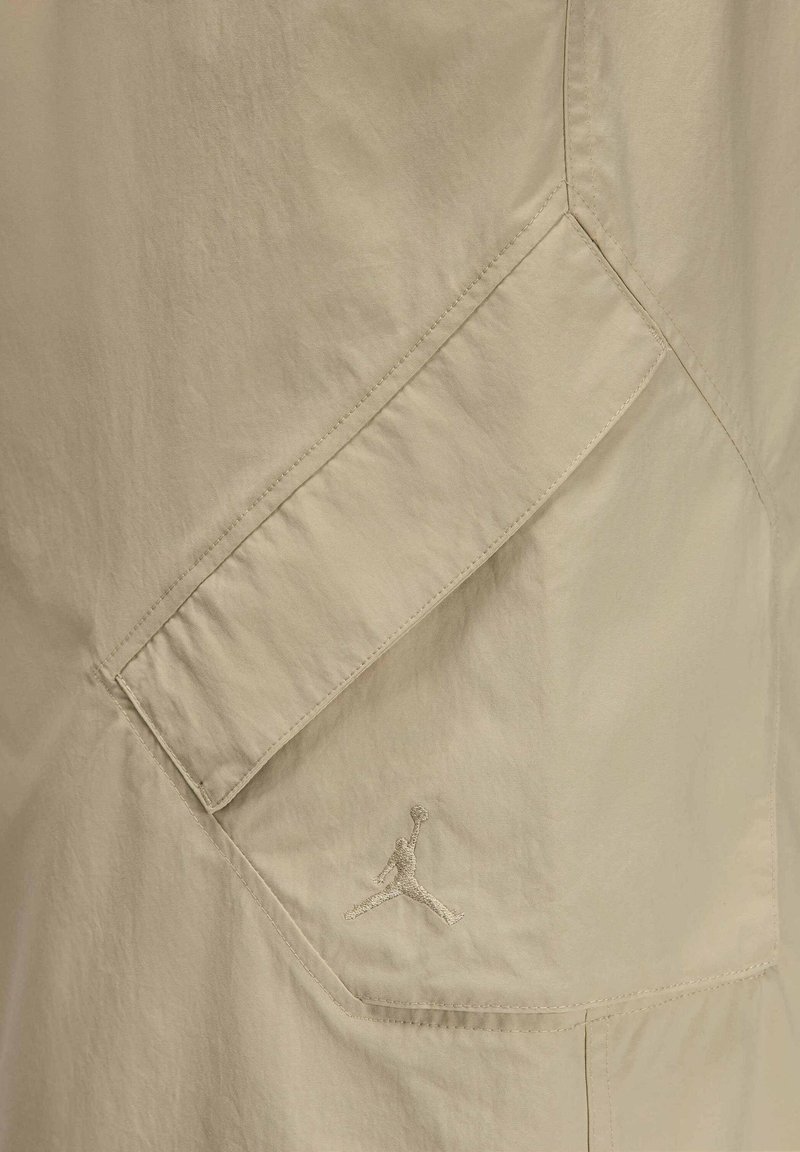 Veste beige avec une poche diagonale et une broderie discrète du logo. Tissu lisse et léger avec une texture nette.