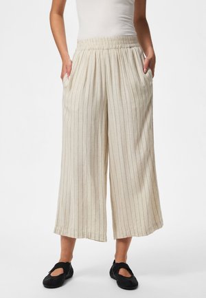 Femme portant un pantalon beige à jambes larges avec fines rayures, taille élastique et chaussures noires slip-on, mains dans les poches, debout sur fond blanc.