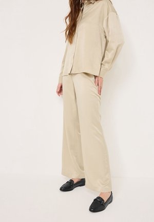 Femme portant une chemise surdimensionnée beige boutonnée et un pantalon large assorti, avec des mocassins en cuir noir, debout devant un fond blanc.