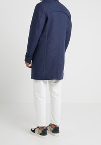 Cappotto in misto lana blu navy con vestibilità su misura, caratterizzato da una spalla a yoke, dettagli con polsini buttonati e linee pulite, abbinato a pantaloni bianchi e sneakers.