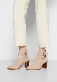 Stivali alla caviglia in suede beige con taglio basso, cintura simile a un'imbracatura, fibbia in tono oro e tacco in legno impilato. Indossati con pantaloni di colore crema.