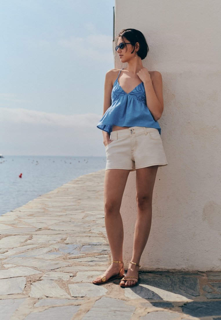 Une femme portant des lunettes de soleil et des vêtements d'été debout contre un mur blanc près d'un chemin en pierre près de la mer.